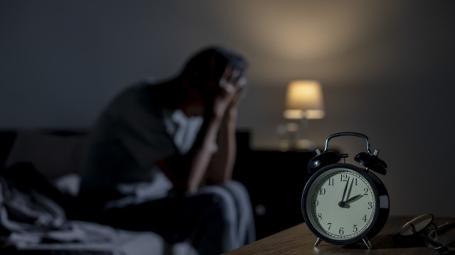 Man sitting on the edge of his bed with his head in his hands. The clock on the nightstand says 2:00 a.m.