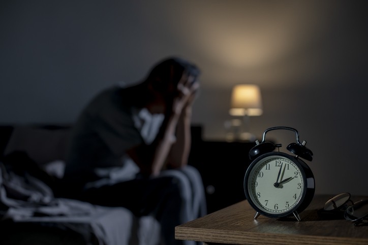 Man sitting on the edge of his bed with his head in his hands. The clock on the nightstand says 2:00 a.m.