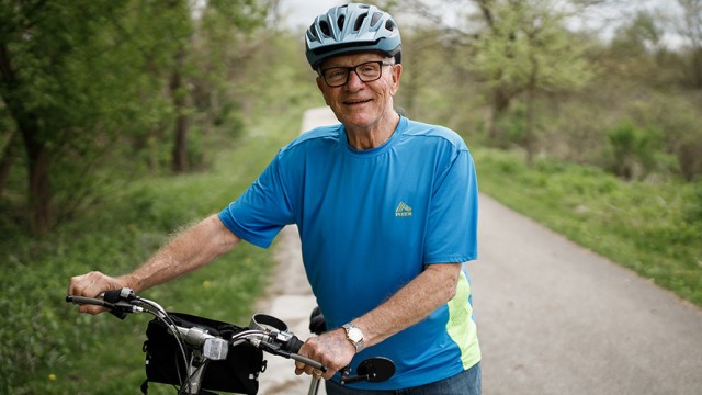 Elderly man with a bike helmet standing beside his bike with trees around