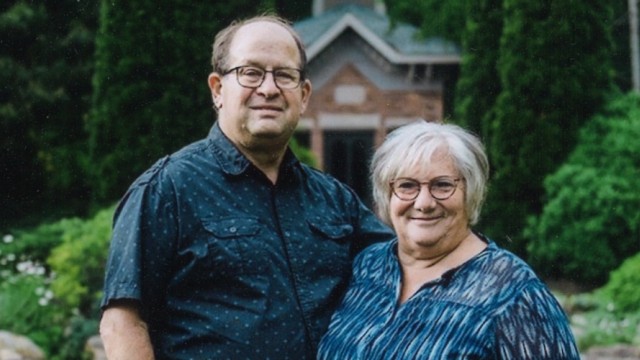 Couple standing shoulder to shoulder with a house and woods in the background
