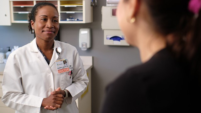 Doctor, standing in an exam room, smiling at a patient.