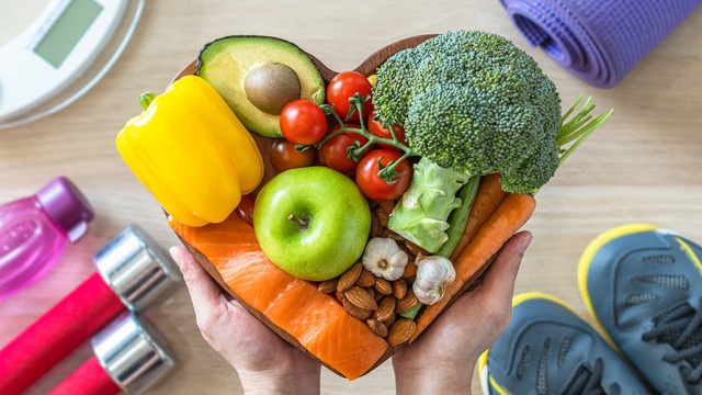 Hands holding healthy foods in the shape of a heart. Exercise equipment is on the floor.