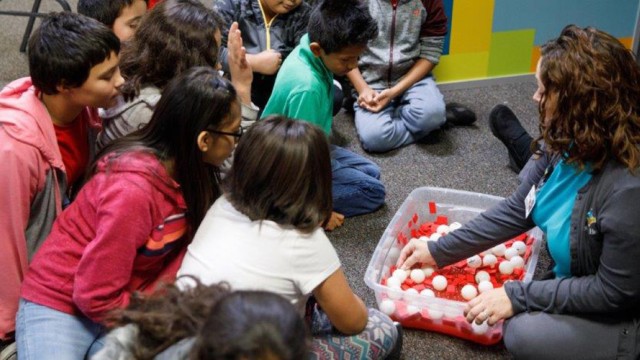 Children sitting on the floor in front of a teacher who is getting items from a tote