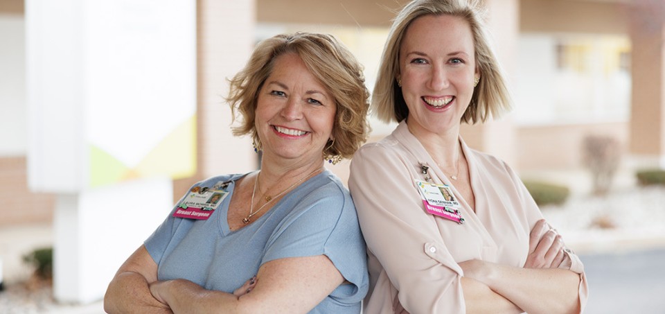 Female breast surgeons smiling while standing back-to-back with arms crossed.