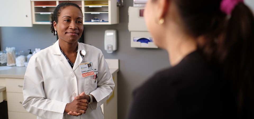Doctor, standing in an exam room, smiling at a patient.