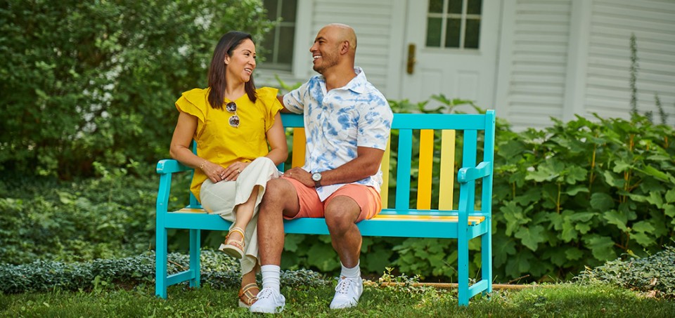Couple talking while sitting on a park bench with flowers and house in the background