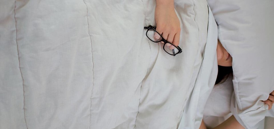 Person lying in bed, holding their glasses while their whole body is under the blanket