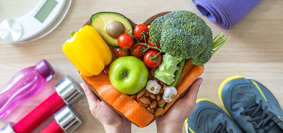 Hands holding healthy foods in the shape of a heart. Exercise equipment is on the floor.