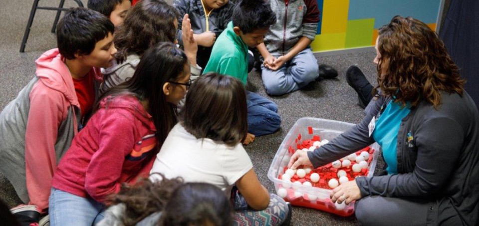 Children sitting on the floor in front of a teacher who is getting items from a tote