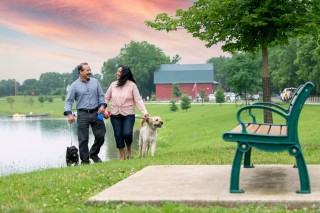 Couple walking their dogs at the park approaching a bench. Water and a barn in the background