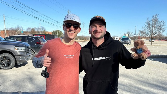 Two men smiling, standing shoulder to shoulder. One man has a thumbs up and one has a teddy bear.