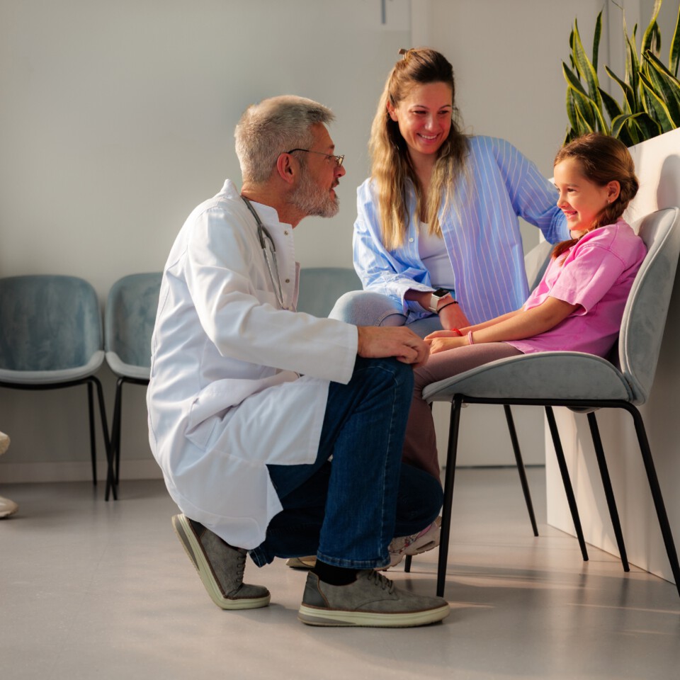 Doctor kneeling in front of a child and her mother sitting in a waiting room
